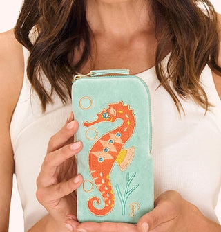 Model holds a rectangular mint green velvet eyeglass case that features colorful embroidery depicting an orange seahorse flanked by seaweed and bubbles