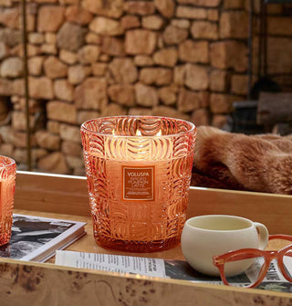 Lit Spiced Pumpkin Latte Voluspa candle staged with coffee mug, reading glasses, book, newspaper, and fur blanked against a stone hearth backdrop
