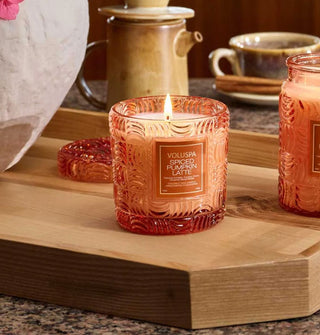 A lit single-wick Spiced Pumpkin Latte Voluspa candle in an embossed orange glass vessel is staged on a wooden tray with another glass candle, its own matching lid placed behind it, and various ceramics
