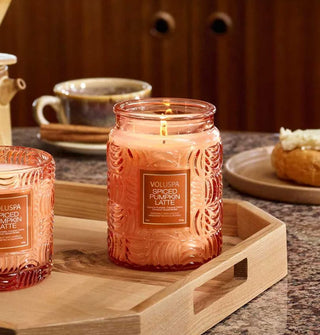 Lit single-wick Spiced Pumpkin Latte in orange embossed glass jar rests on a wooden handled tray with another Voluspa candle of the same scent and glass style and a coffee mug and bagel in the background