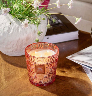 Lit three-wick Spiced Pumpkin Latte Voluspa candle in embossed glass vessel on a wooden tabletop with potted flowers and books
