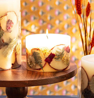 Lit single-wick short pillar candle with embedded apple slices, flowers, and ferns is staged on a wooden pedestal with other, larger candles like it against a yellow floral wallpapered backdrop