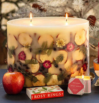 Large lit two-wick statement candle features apple slices, ferns, flowers, cinnamon sticks, and berries embedded in its wax and is staged with an apple, Rosy Rings matchbox, and small lit tin candle against a backdrop of snowy coniferous branches and pine cones