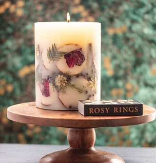 Lit single-wick pillar candle with embedded apple slices, ferns, and other botanicals is staged on a wooden pedestal with Rosy Rings matchbox