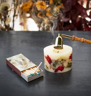 Smoke rises from a freshly snuffed stout pillar candle embedded with apple slices, flower petals, and ferns staged next to an opened box of matches on a wooden tabletop