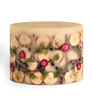 Large two-wick candle features apple slices, small ferns, red and white flowers, cinnamon sticks, and berries embedded in its wax