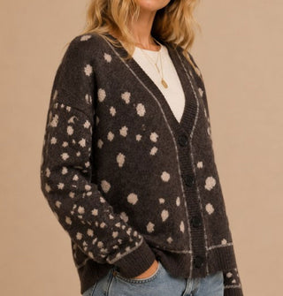 Model with hand in pocket wears a brown V-neck cardigan with light spots all over and a thin light line along the neckline, button placket, and sleeve cuffs