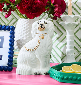 White ceramic Staffordshire dog statue on a pink shelf staged with other ceramic home goods against a white and green patterned backdrop