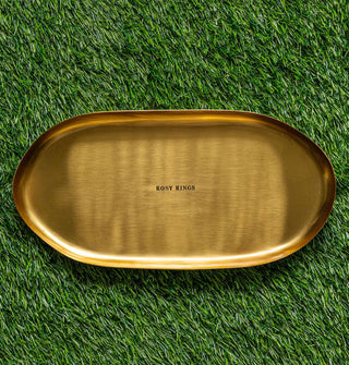Oblong gold-toned platter with shallow lip and "Rosy Rings" stamped in the center rests on green astroturf