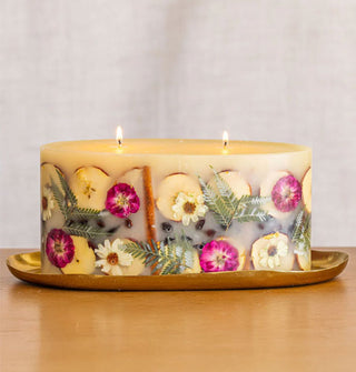 A lit two-wick oblong candle with embedded apple slices, ferns, flowers, cinnamon sticks, and berries rests on a slightly irregularly shaped gold platter