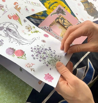 Model's hands remove a sticker from a sheet of floral designs surrounded by other decorative papers