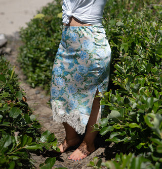 Model standing among shrubbery wears a satin pencil skirt with lace trim and blue floral print