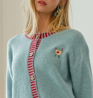 Model wears a powder blue cardigan with red striped button placket and neckline and a small embroidered rooster on the left chest