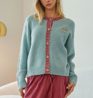 Model wears a powder blue cardigan with red striped button placket and neckline and a small embroidered rooster on the left chest