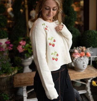 Model wears a white cardigan with strawberry embroidery against a backdrop of potted plants and a bench