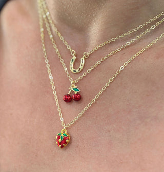 Model wears three gold necklaces of varying lengths, one with a horseshoe charm, one with a red cherries charm and one with a red strawberry charm