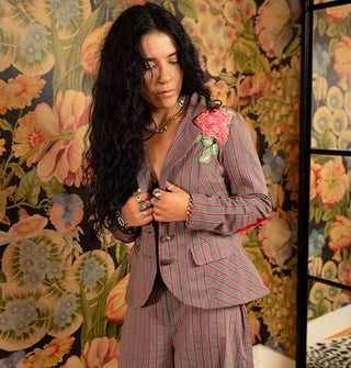 Model wears a gray and red striped blazer with rose shoulder appliqué paired with matching pants against a bold floral backdrop