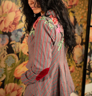 Model wears a gray and red striped blazer with embroidered floral shoulder appliqués and red velvet elbow patch against a bold wallpapered backdrop