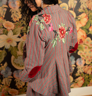 Model wears a gray and red striped blazer with embroidered floral shoulder appliqués and red velvet elbow patches against a bold wallpapered backdrop