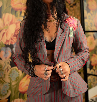 Model wears a gray and red striped blazer with rose shoulder appliqué paired with matching pants against a bold floral backdrop