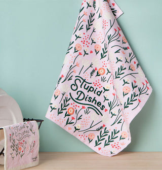 A white cloth that features whimsical floral patterning and the words, "Stupid Dishes" embroidered in dark green hangs against a mint green backdrop near a drying rack slung with a dish cloth of the same style