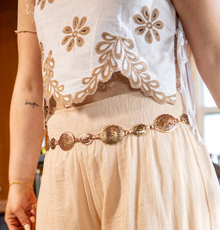 Model wears a white sleeveless top with ties at the sides and brown floral eyelet embroidery with a pair of stretch-waist linen pants accented with a gold belt