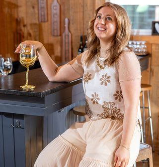 Smiling model sitting at a bar wears a white sleeveless top with ties at the sides and brown floral eyelet embroidery