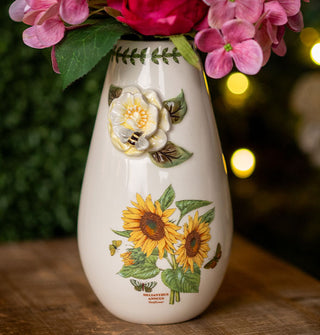 White teardrop-shaped vase features sunflowers, a white gardenia, butterflies, a bee, and a vine border around the opening, and holds a bouquet of pink flowers