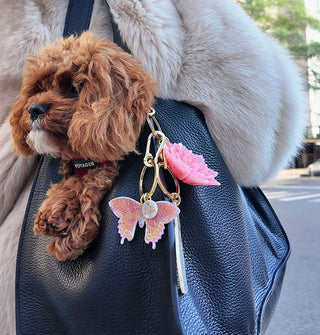 Model in fur coat holds a pebbled black leather bag over shoulder with a brown and white, curly-haired dog inside and pink swallowtail butterfly and pink flower keychains attached to its strap