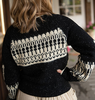 Model with back to camera wears a black sweater with an intricate white intarsia design on the back and around the lower sleeves