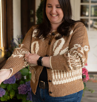 Smiling model wears and pulls up the sleeve of a brown button-down cardigan with swans, bows, and intricate border designs, and white edging along the neckline and sleeve cuffs