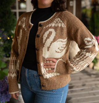 Model wears a brown button-down cardigan with swans, bows, and intricate border designs, and white edging along the neckline and sleeve cuffs