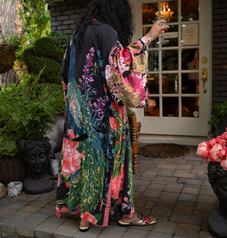 Model holding crystal goblet and turning away from camera wears an ankle-length black robe with ultra-wide sleeves and an all-over print of large, colorful flowers and peacocks paired with metallic floral mules and fishnets