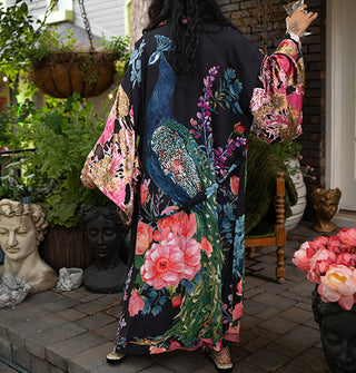 Model holding a crystal goblet and with back to the camera wears an ankle-length black robe with ultra-wide sleeves and an all-over print of large, colorful flowers surrounding a central peacock