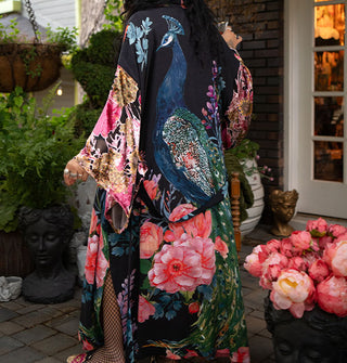 Model with back to the camera wears an ankle-length black robe with ultra-wide sleeves and an all-over print of large, colorful flowers surrounding a central peacock