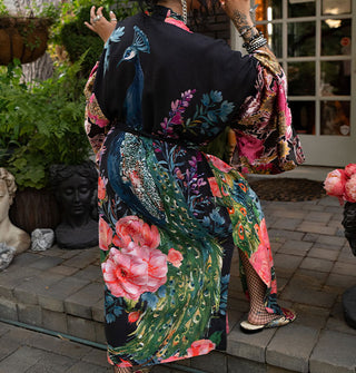 Model with back to the camera wears an ankle-length black belted robe with ultra-wide sleeves, side slit, and an all-over print of large, colorful flowers surrounding a central peacock