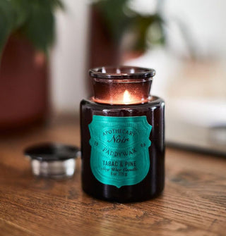 A lit Apothecary Noir Paddywax Tabac & Pine candle shines from inside a black apothecary-style jar on a wooden surface with houseplants in the background