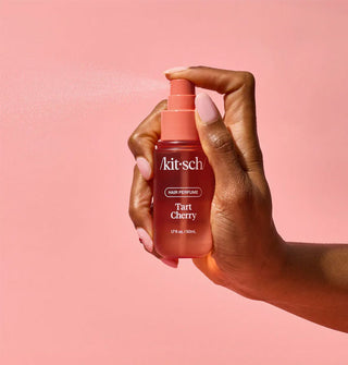 Model's hand dispenses a fine mist of Kitsch Tart Cherry Hair Perfume against a light pink backdrop