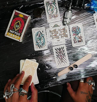 Model wearing silver rings arranges Tattoo Tarot mini cards on a plastic-wrapped surface staged with tattoo gun and ink pots