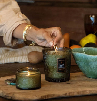 Model lights a Temple Moss candle in a green embossed glass vessel alongside a smaller one like it on a rustic wooden tabletop staged with a ceramic bowl and produce
