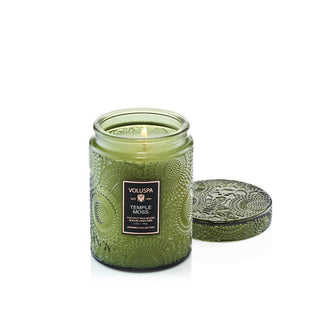 Lit single-wick Temple Moss Voluspa candle in an embossed green glass vessel with matching lid removed and set to the side