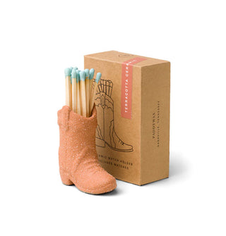 A terracotta-colored cowboy boot figurine holding several wooden matchsticks with light blue tips sits next to its brown box packaging