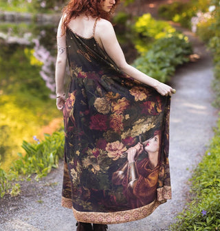 Model wears a long sleeveless dress with portrait of Joan of Arc among lush florals on the skirt with contrasting stained glass patterned border
