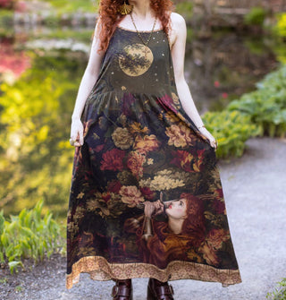 Model wears a long sleeveless dress with portrait of Joan of Arc among lush florals on the skirt, a full moon partially obscured by a tree branch on the bodice, and a contrasting stained glass patterned border