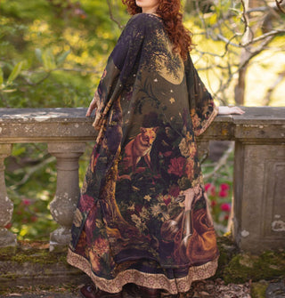 Model standing at a balustrade with back to the camera wears a long, kimono-style robe featuring a nighttime woodland motif with flowers, a fox, and a sword-wielding figure at the bottom along a contrasting printed border