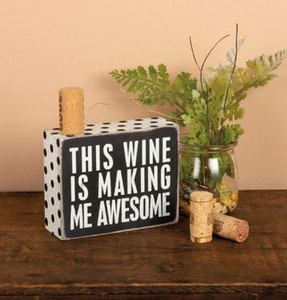 Black and white "This Wine Is Making Me Awesome" wooden box sign is staged with wine corks and a jar of ferns and twigs on a rustic wooden tabletop