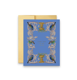 Blue greeting card in front of a gold envelope is adorned with a border of intricate vines perched upon by vibrantly colored parrots, inside of which are the words, "Thou art on my mind" in small gold lettering