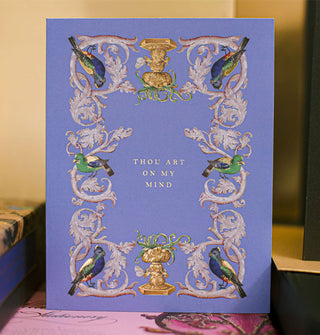 Blueish-purple greeting card is adorned with a border of intricate vines perched upon by vibrantly colored parrots, inside of which are the words, "Thou art on my mind" in small gold lettering