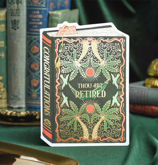 Green greeting card with gold-accented palm trees, sun, birds, and hammock designs made to look like a book cover with the words, "Thou Art Retired" printed on the front in gold and, "Congratulations" printed on the spine