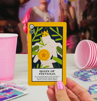 Model's hand holds a Queen of Pentacles card up against a blurred backdrop of other cards, pink cups, and partygoers